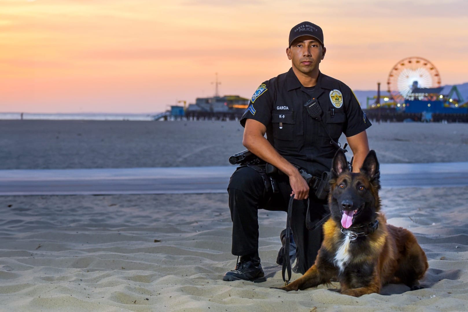 Police officer in uniform with a K9 dog on a sandy beach at sunset, pier and Ferris wheel in the background.