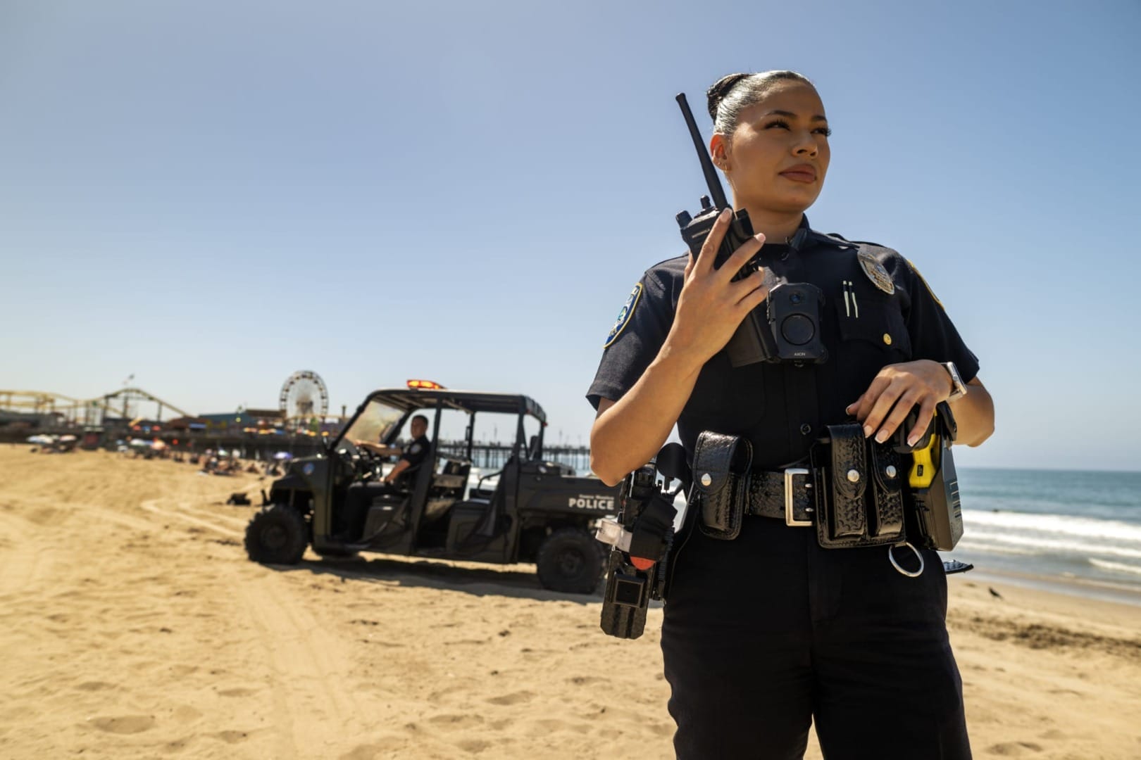 Police officer on a beach holding a radio, with a patrol vehicle and people in the background near the ocean.