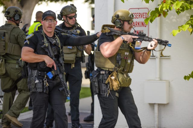 Police officers in tactical gear holding rifles, moving in formation near a building with a reserved parking sign.