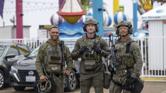 Three uniformed officers in tactical gear stand outdoors in front of amusement park rides and parked cars.