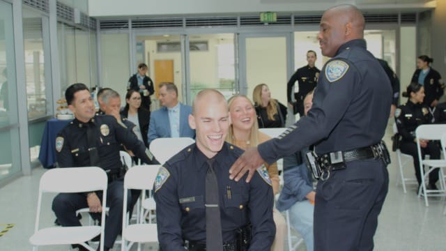 Police officer congratulating a fellow officer seated in a room with several other people during a formal event.