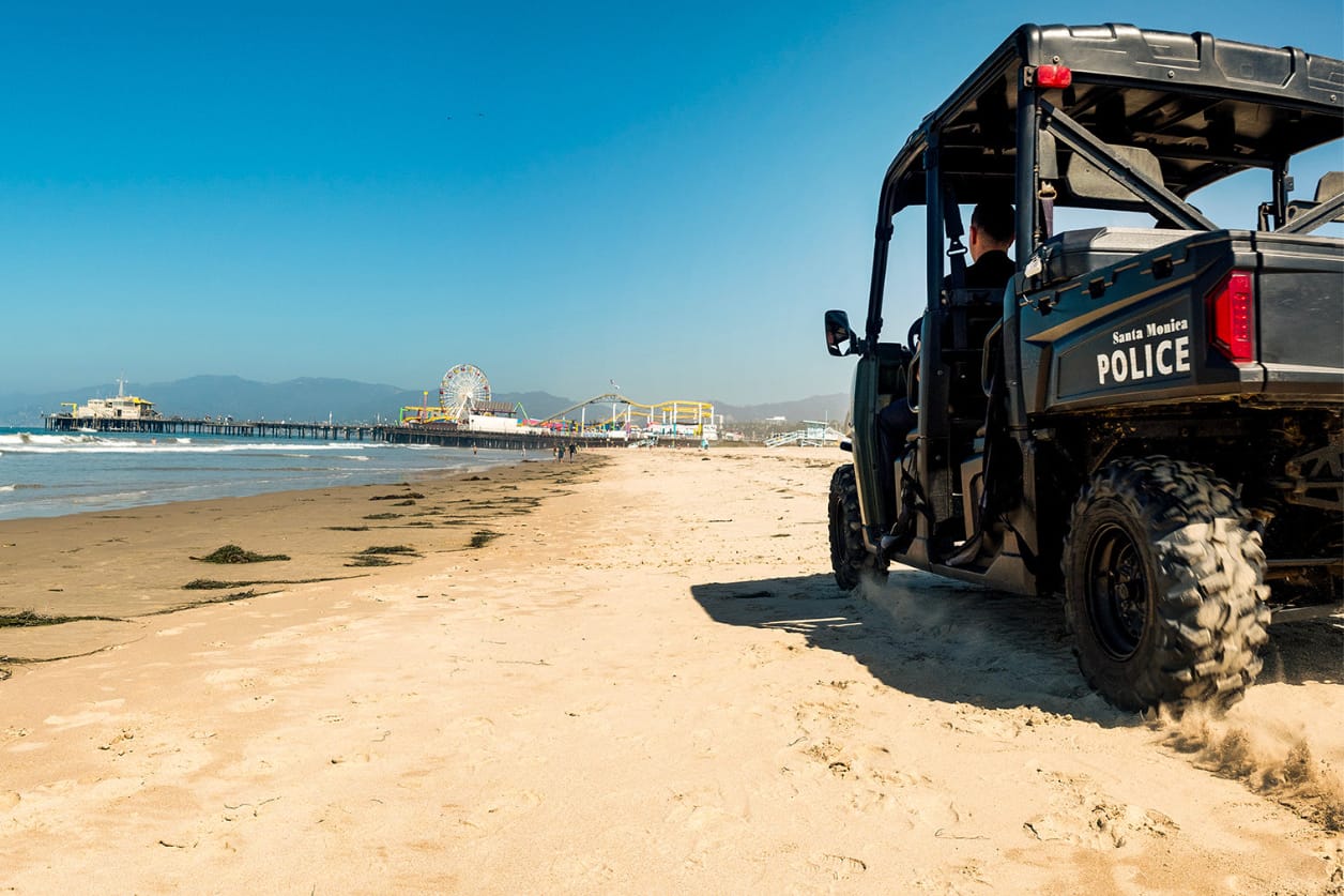 Beach patrol vehicle driving along sandy shore near pier on a clear day.