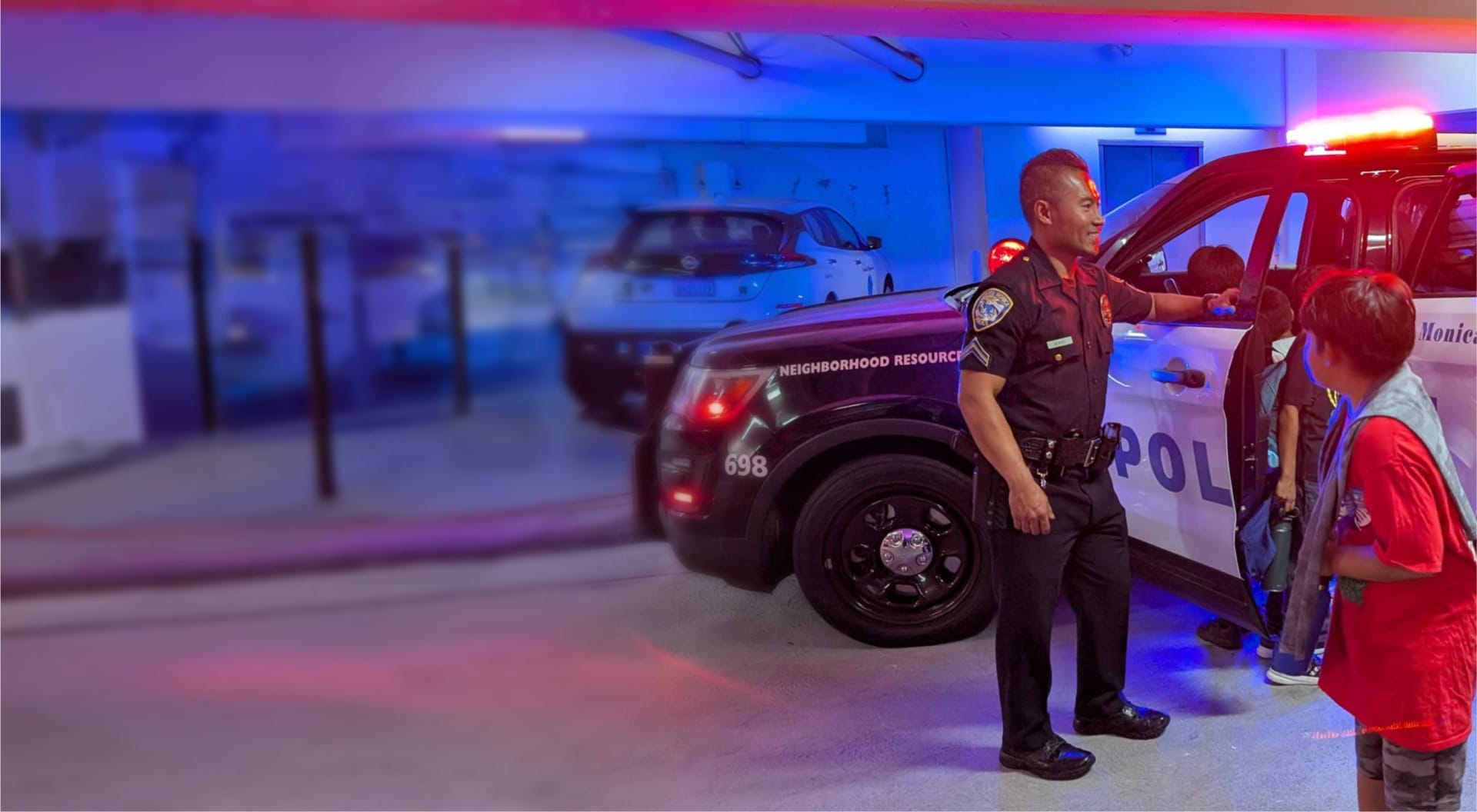 Police officer talking to group of children beside patrol car in parking garage with blue and red lights.