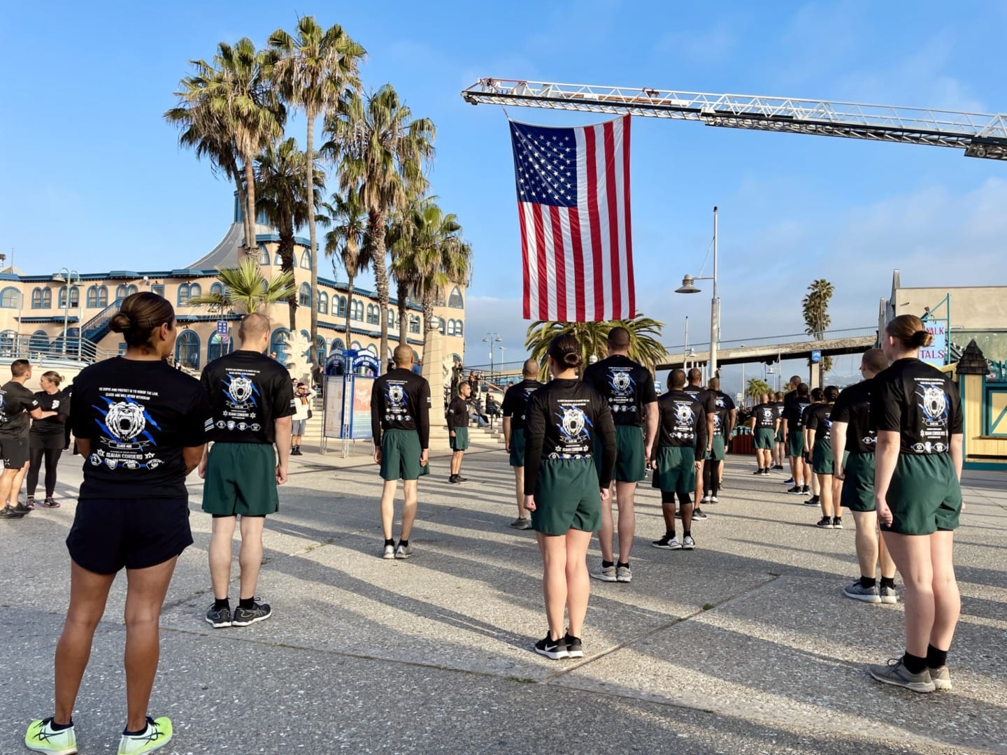 People in uniform standing in formation under a large American flag outdoors on a sunny day with palm trees in the background.