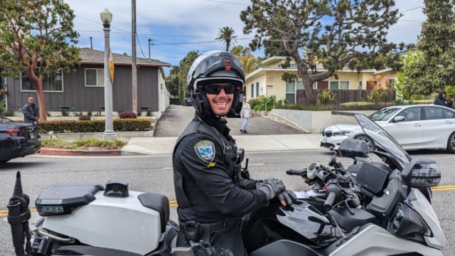 Police officer on a motorcycle, wearing a helmet and smiling, with residential houses and trees in the background.