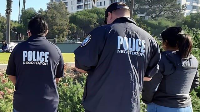 Three police negotiators standing outdoors facing away, wearing jackets labeled "Police Negotiator" in a green park setting.