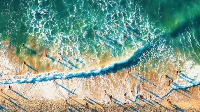 Aerial view of a busy beach with people sunbathing and playing near ocean waves, lifeguard tower and umbrella visible.
