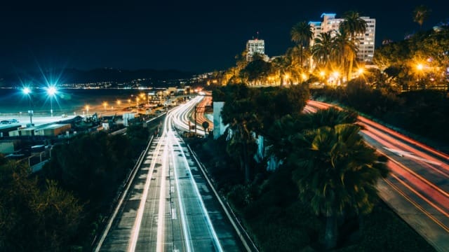 Night view of a coastal city with light trails from traffic, illuminated buildings, and palm trees under a dark sky.