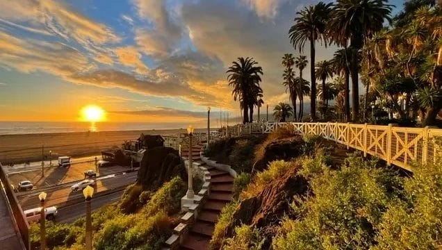 Sunset over a beach with palm trees and a pathway, vibrant clouds in the sky, and a beachside road.