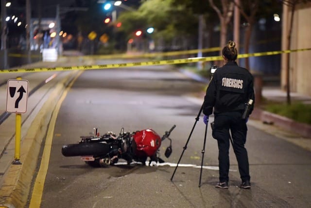 Forensic investigator photographing a motorcycle accident scene at night with yellow police tape in the background.