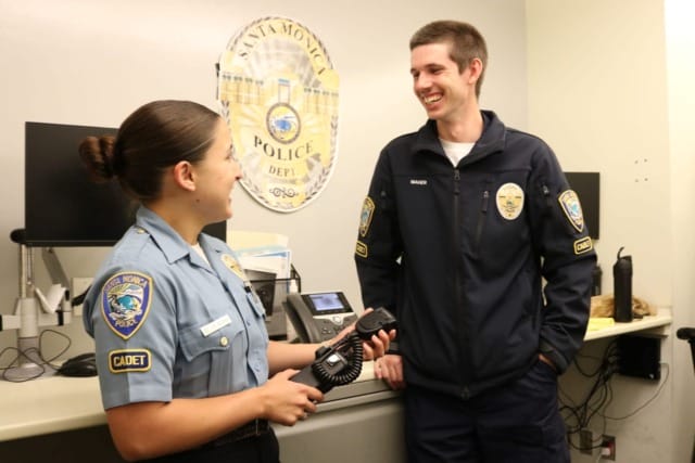 Two police officers in uniform talking and smiling inside a police department office next to a desk and computer.