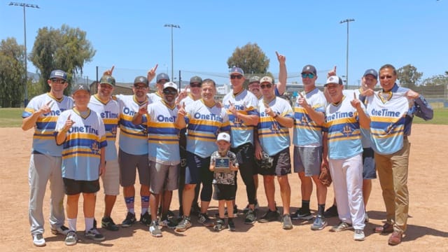 Men and a child in matching baseball jerseys pose on a field, holding up one finger in a celebratory gesture.