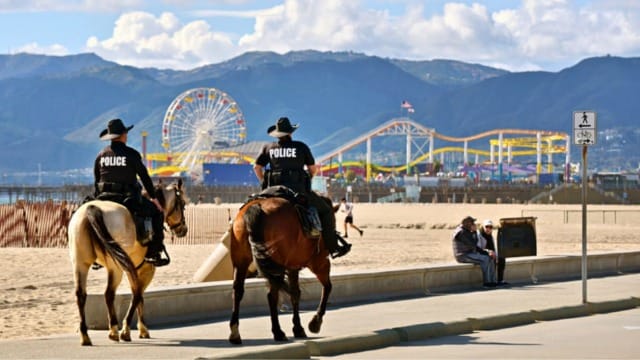 Mounted police patrolling beach area with amusement park and mountains in the background under a partly cloudy sky.
