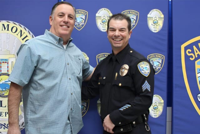 Two men smiling, one in a police uniform, standing in front of Santa Monica Police Department banners.