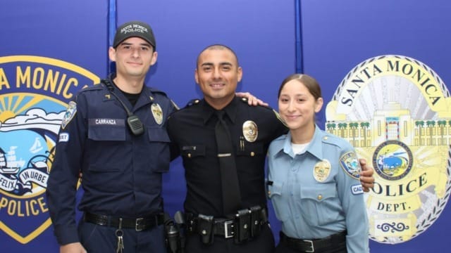 Three police officers smiling in uniform, standing in front of Santa Monica Police Department banners.
