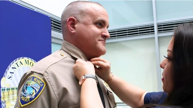Person adjusting a police officer's uniform next to Santa Monica Police badge in an indoor setting.
