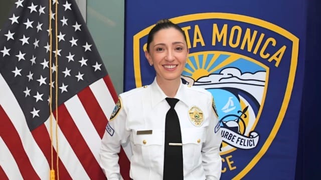 Uniformed police officer smiling in front of Santa Monica Police banner and American flag.
