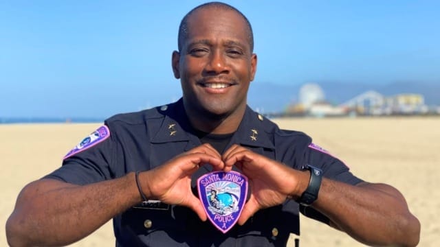 Police officer smiling on a beach, holding a Santa Monica Police patch shaped like a heart in front of him.