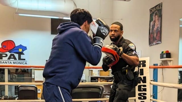 Police officer and young man sparring in a boxing ring at a gym, with exercise equipment in the background.