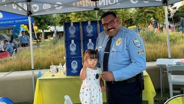 Police officer smiles with a young girl at a community event under a tent with JOIN SMPD banner, colorful toys on table.