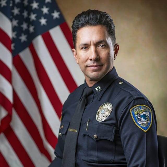 Police officer in uniform seated in front of the American flag, displaying badges and insignia.