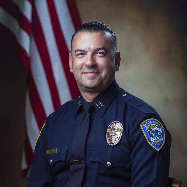 Uniformed police officer sitting in front of an American flag backdrop, smiling, in a formal portrait setting.