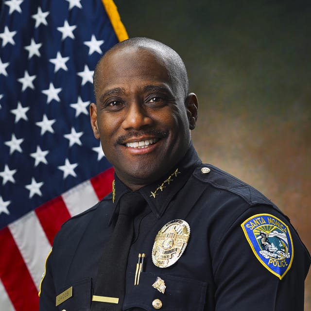 Police officer in uniform smiling with a flag in the background.
