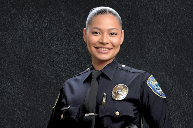 Smiling police officer standing confidently in uniform against a dark background.