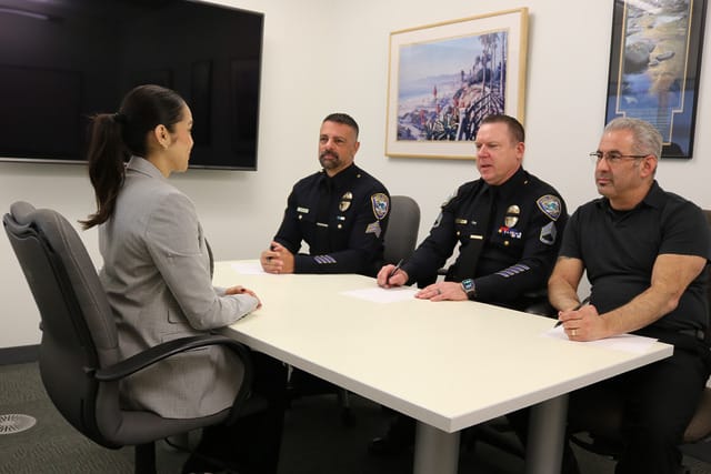 Person in a business suit sits across from three people in uniform and a civilian at a conference table in an office setting.