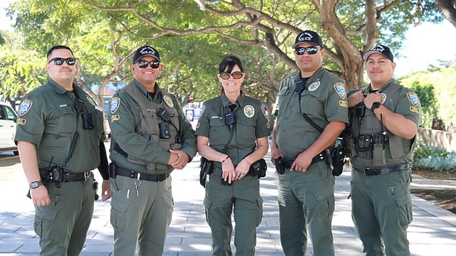 Group of security personnel in uniform standing on a paved walkway with trees in the background during the day.
