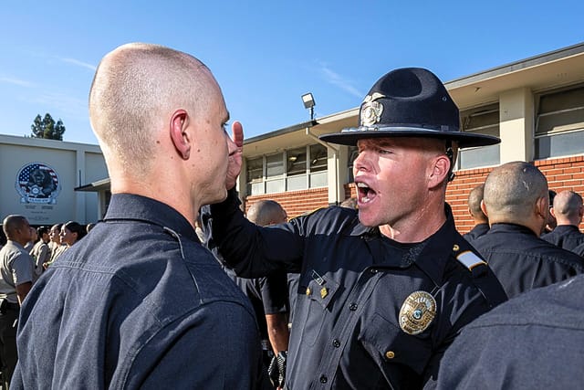Police officer in uniform energetically instructs a trainee outside a brick building in a group training session.