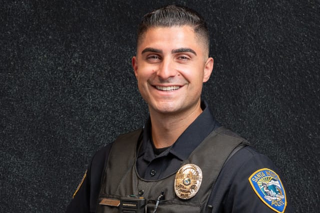 Smiling police officer in uniform and tactical vest, standing with hands on belt against a dark textured background