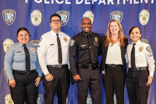 Five police department personnel smiling in uniform in front of a banner with the department's name and emblem.